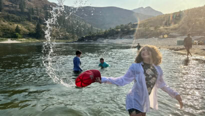 A girl splashes on the shore of a river with people frolicking in the shallow water in the background