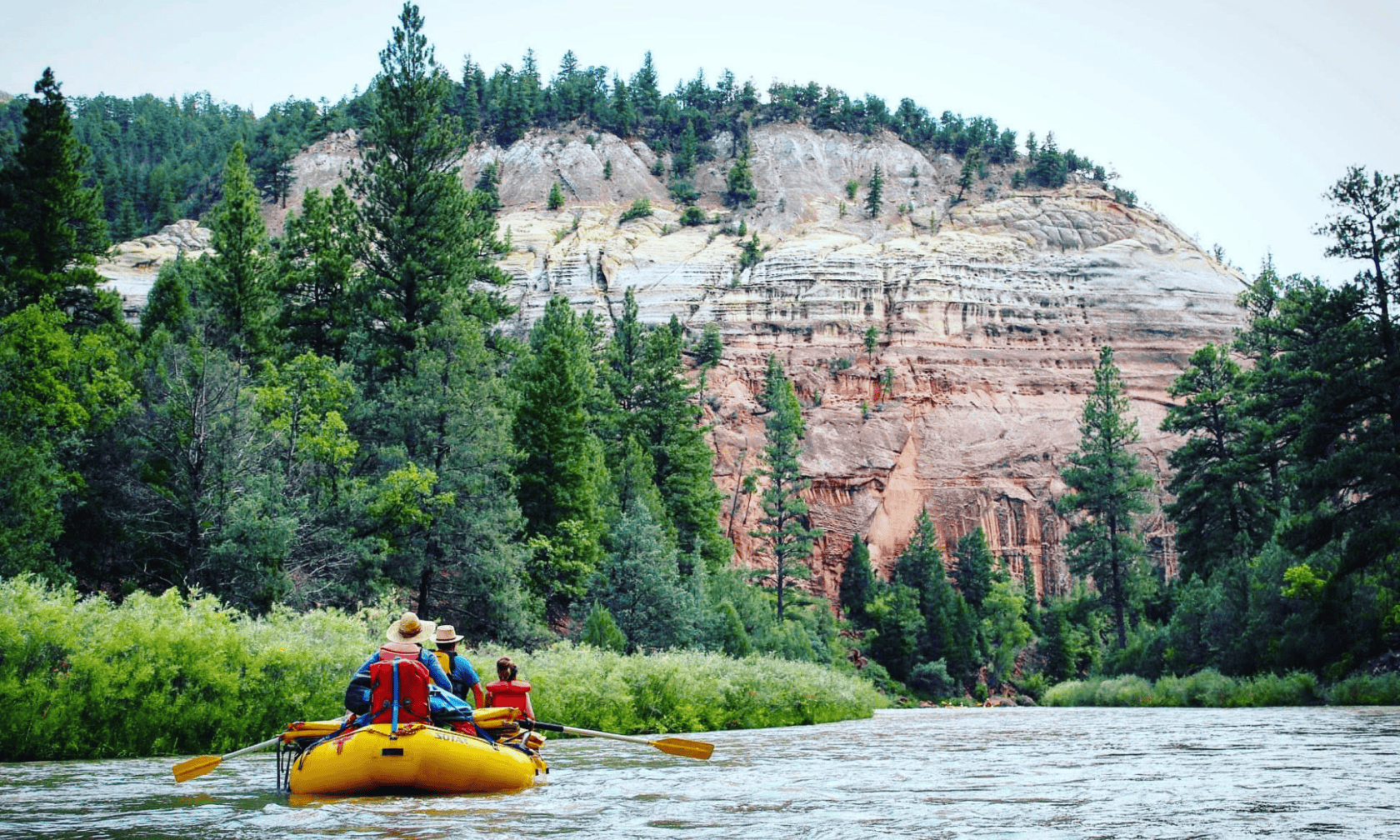 A raft floats through a pine-dotted river canyon with stunning red walls on the Rio Chama, a popular New Mexico whitewater trip