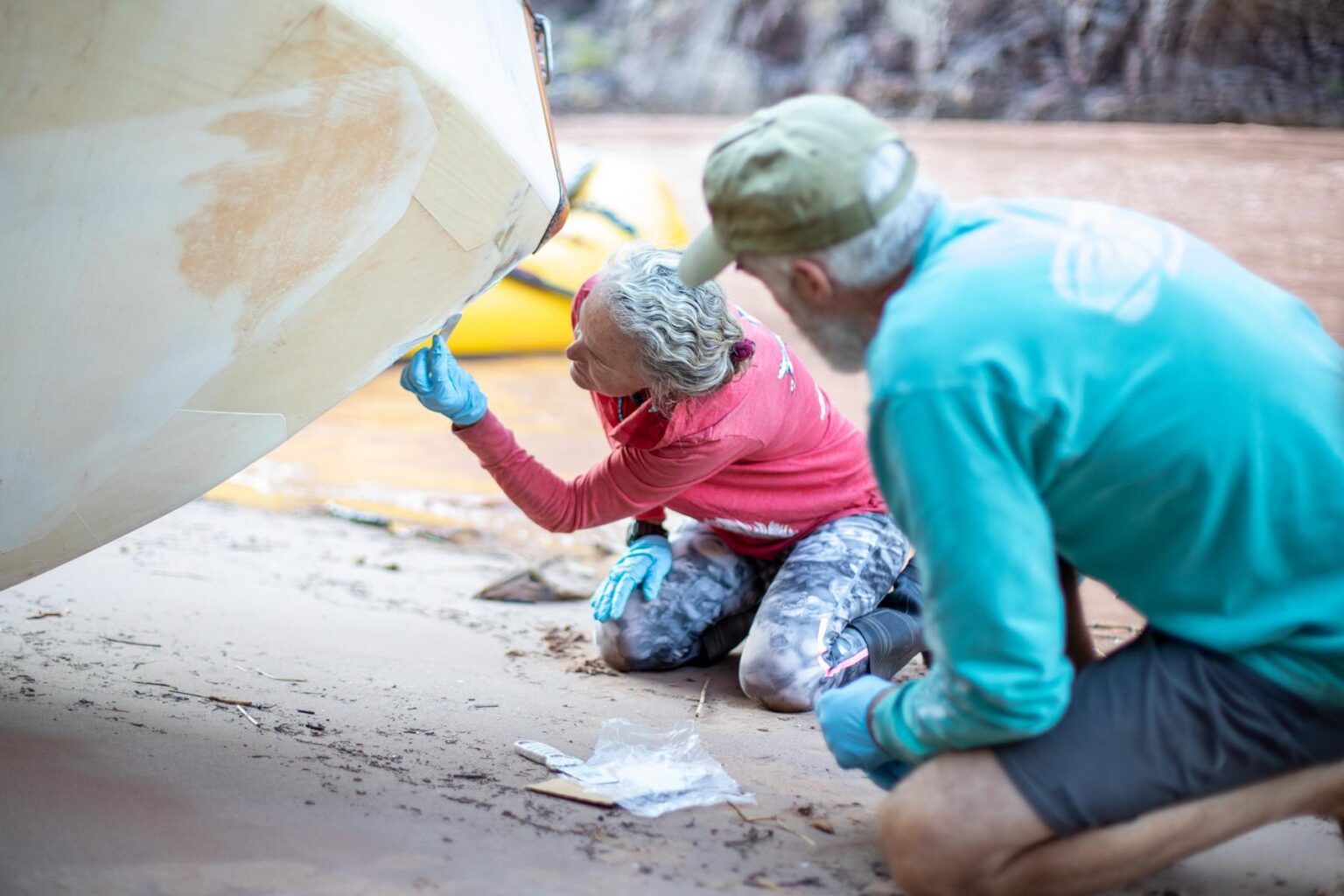 A woman and a man patch the bottom of a boat that's pulled up on shore alongside the Colorado River.