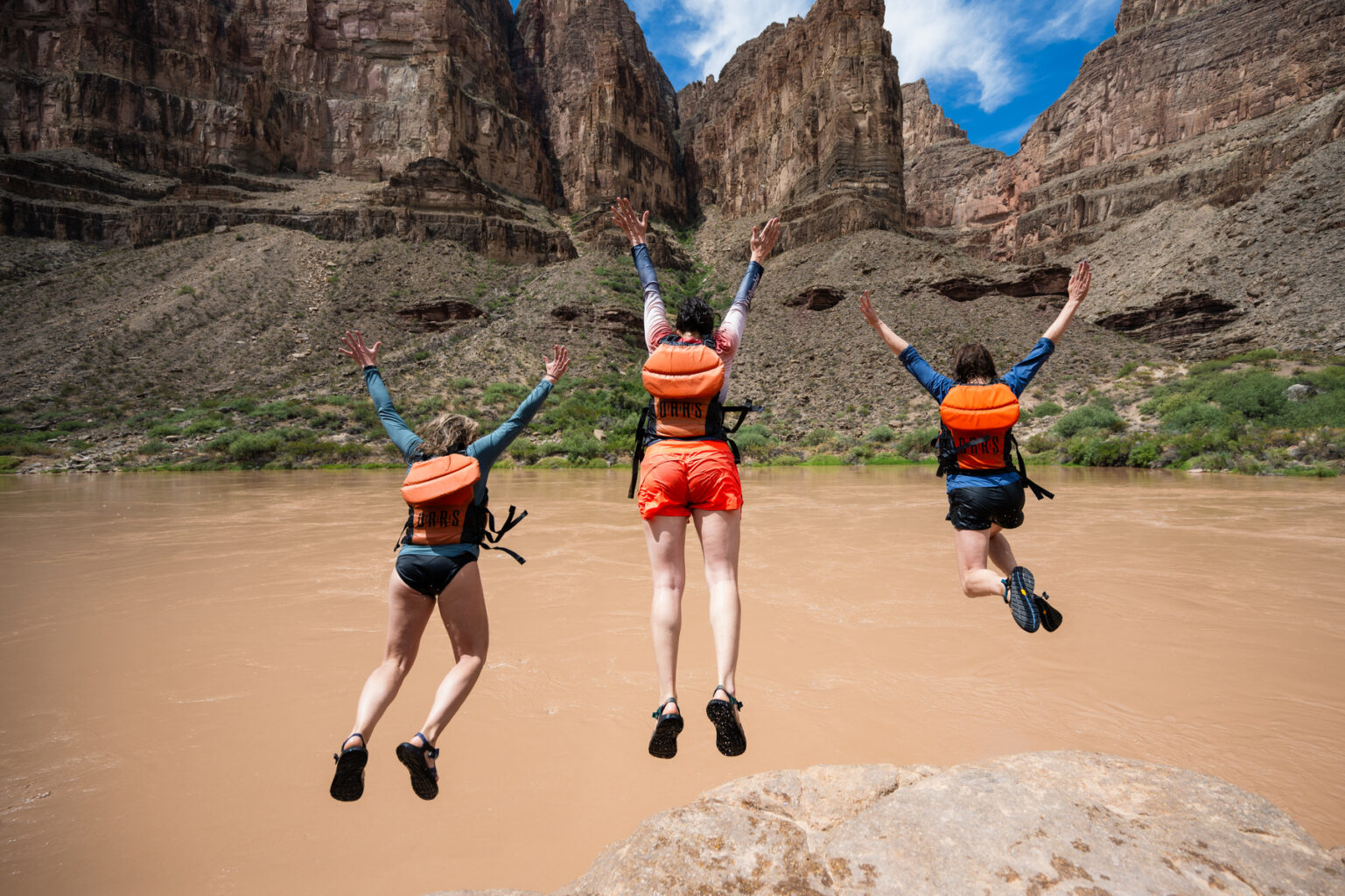 Three women in PFDs jump into a brown and silty Colorado River in Grand Canyon