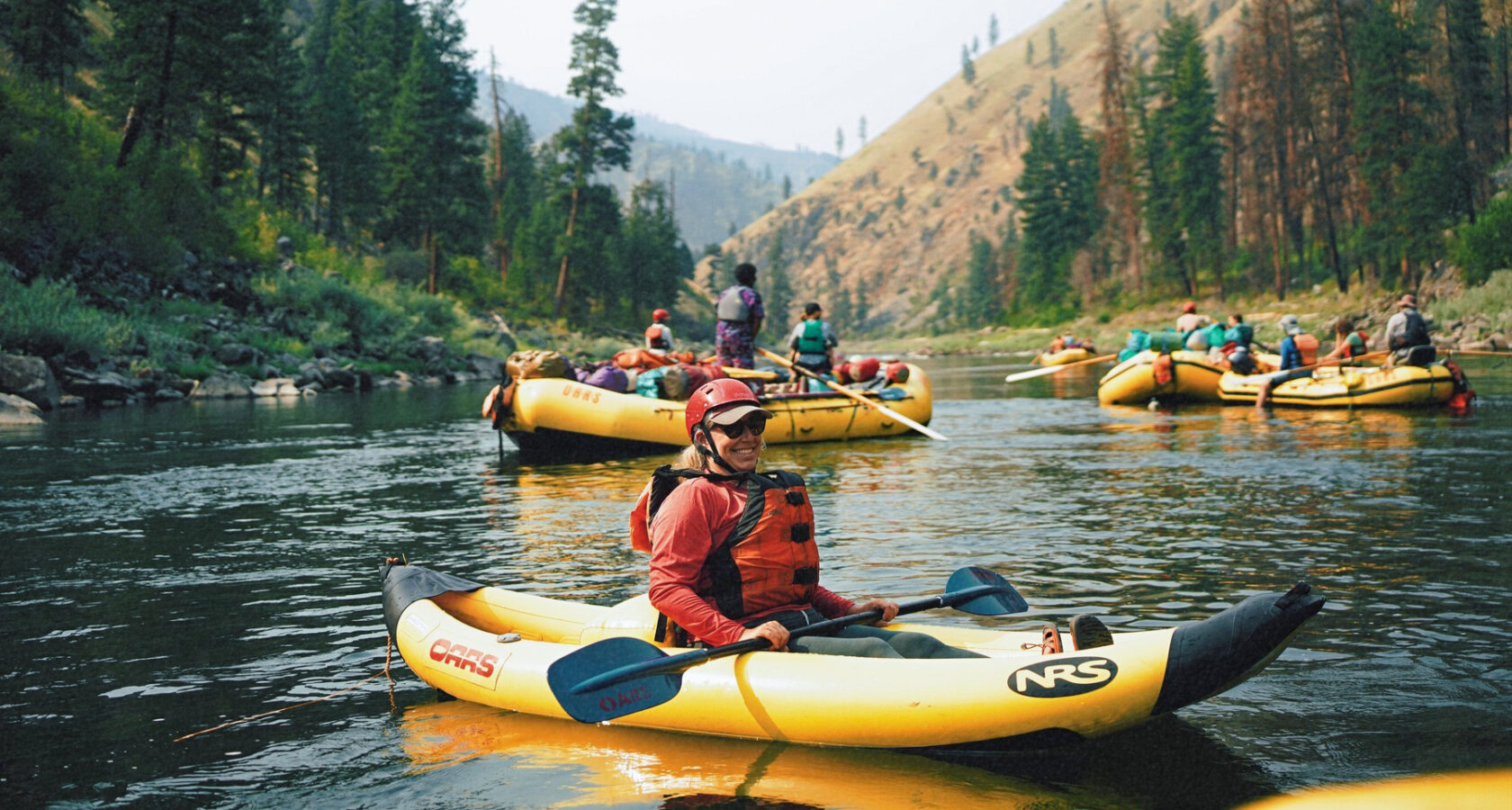 A woman wearing a PFD and helmet sits in an inflatable kayak amongst a group of yellow rafts on the Main Salmon River
