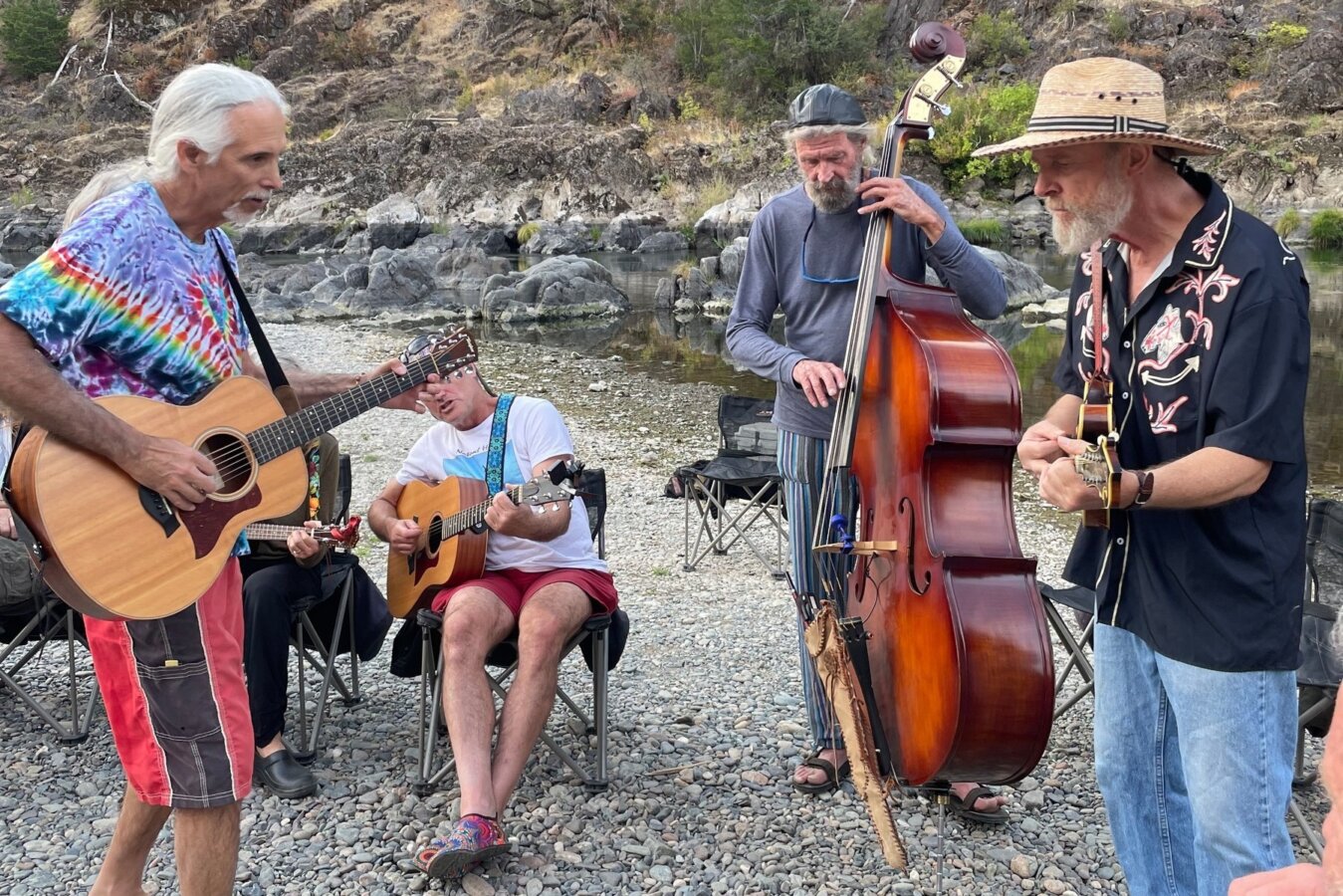 A group of musicians play acoustic instruments on a rocky beach on the river.