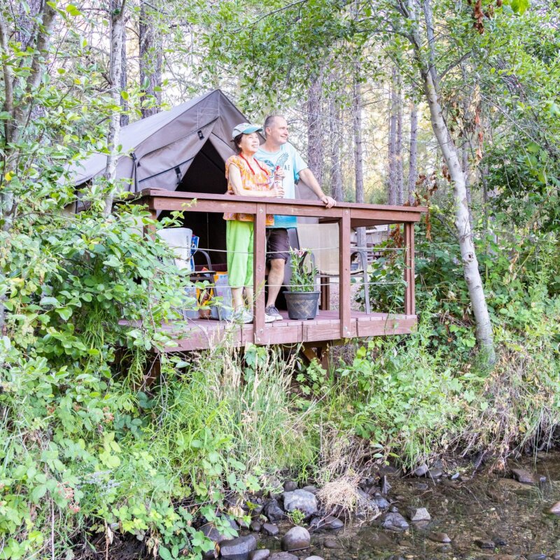 Two people stand on the deck of their canvas tent cabin and look out at the American River which runs right by the cabin.