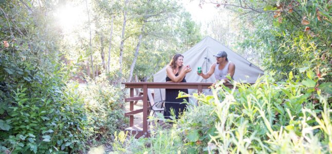 Two people stand on the deck of a canvas tent cabin and cheers their soda cans.